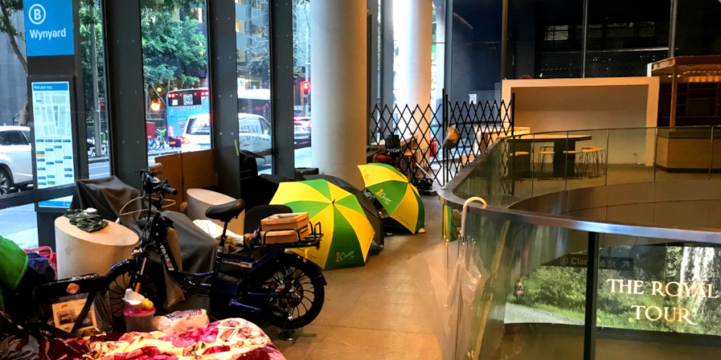 Interior view of a city space with seating and a glass railing, featuring a makeshift living area with a bicycle, blankets, and brightly coloured umbrellas.