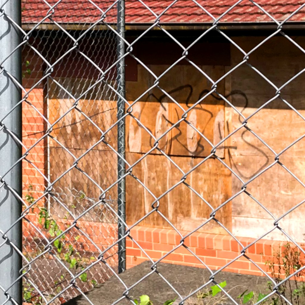 A view through a chain-link fence showing a boarded-up building with graffiti on the wooden panels.