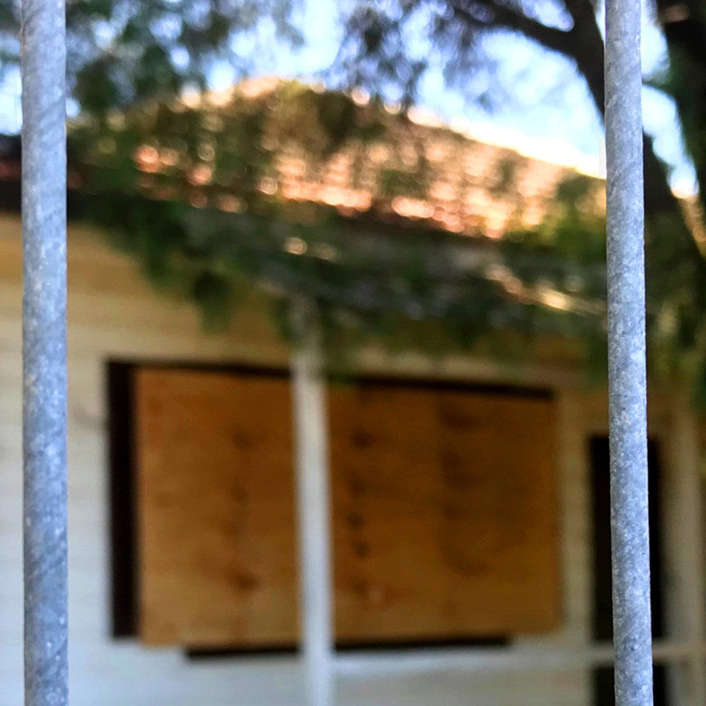 A blurred view of a house with boarded windows, seen through metal bars.