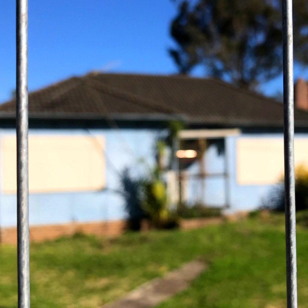 A partially obscured view of a blue house with boarded windows, seen through metal fencing, set in a grassy area under a clear blue sky.