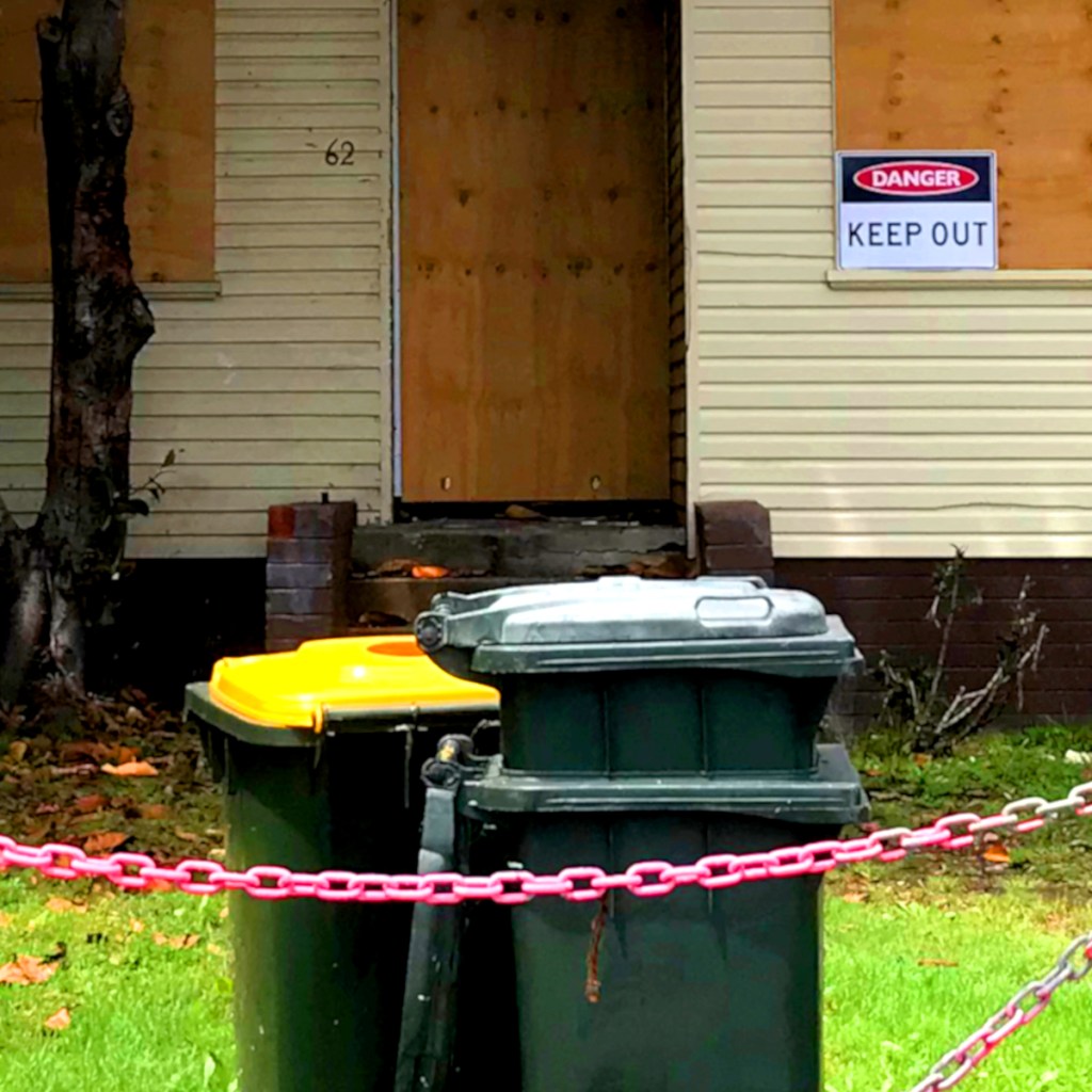 A pair of rubbish bins, one yellow and one green, placed near a boarded-up house. A sign on the wall reads 'DANGER KEEP OUT'.