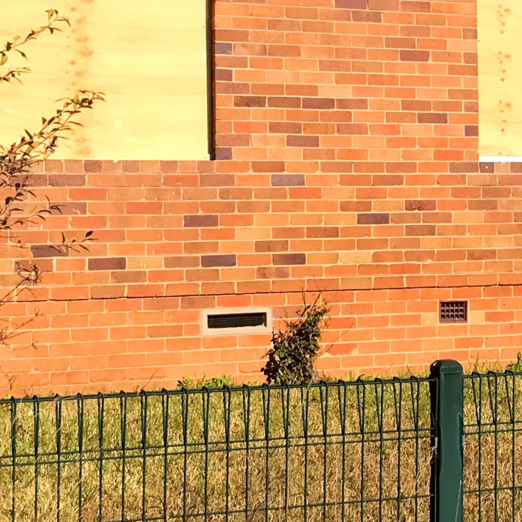 A section of a brick wall featuring a vent at the bottom, surrounded by a patch of grass and a green metal fence.