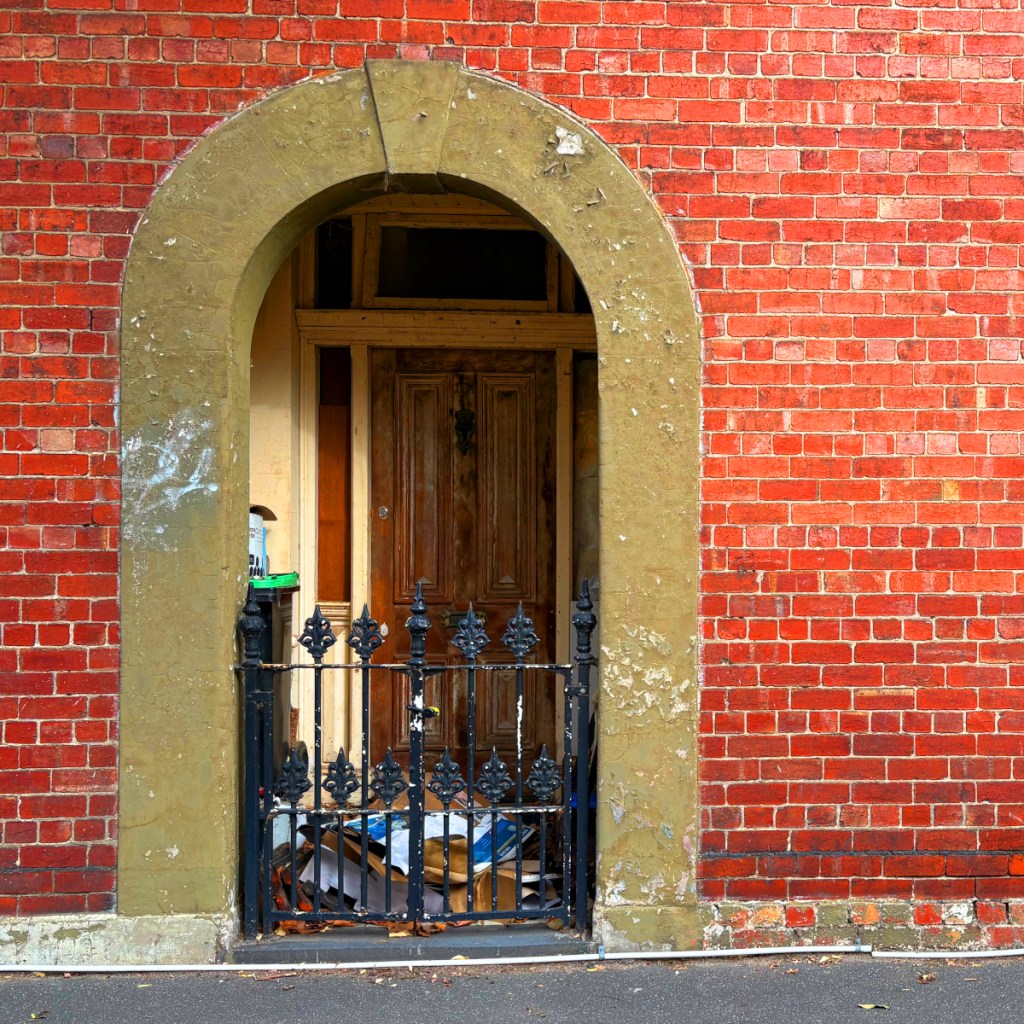 An ornate archway with a wooden door and a decorative black wrought iron gate, set against a red brick wall.
