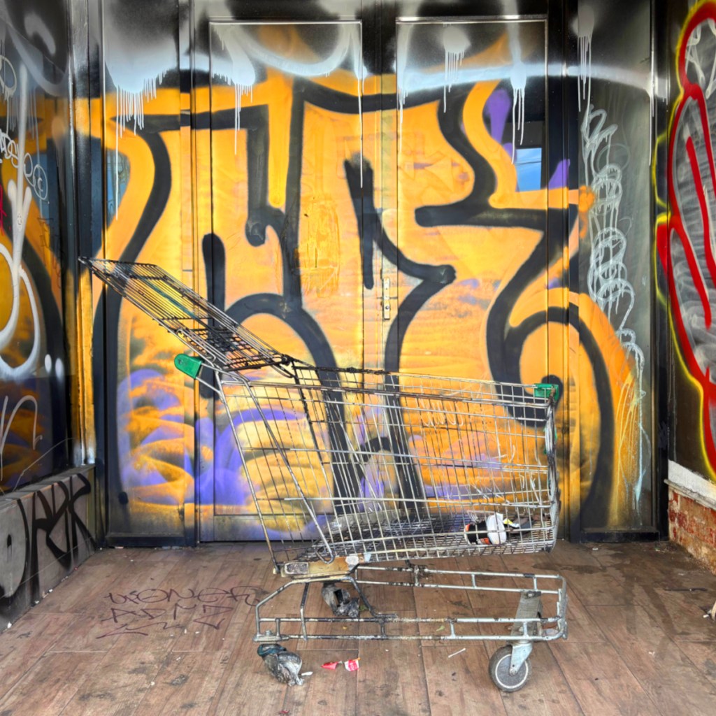 An abandoned shopping trolley placed in front of a vibrant graffiti wall featuring orange and purple designs.