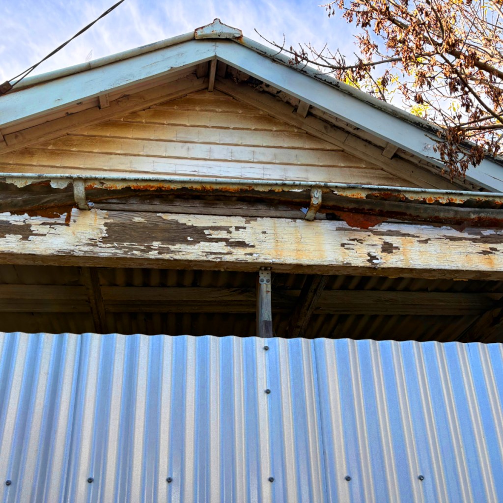 Close-up view of the deteriorating gable of an old building, showcasing peeling paint and weathered wood, with a corrugated metal fence in the foreground.