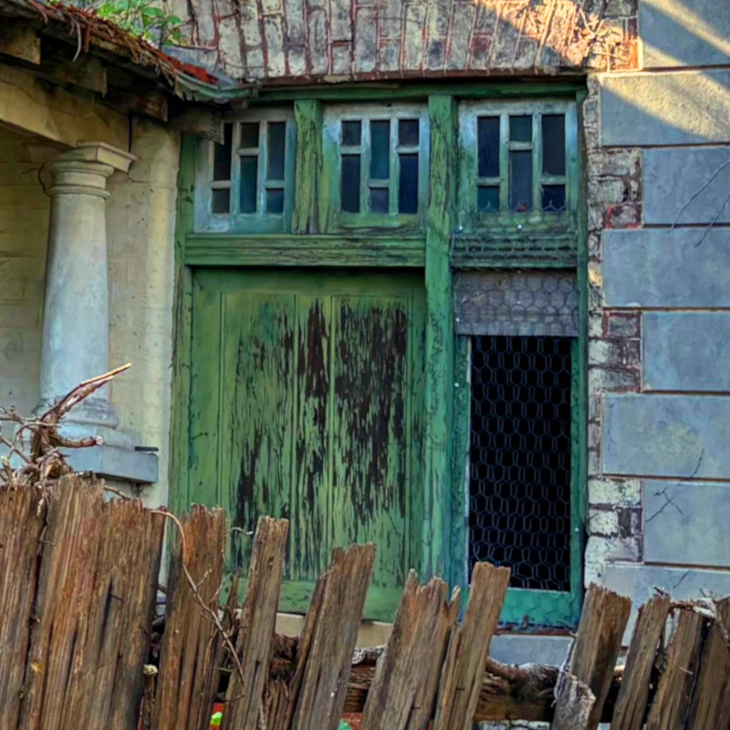 An old, weathered green door with a window, set against a brick wall, partially obscured by a wooden fence.