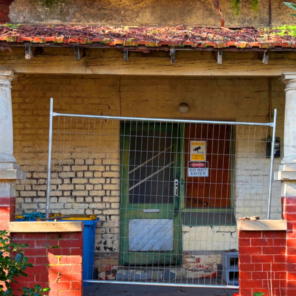 Front entrance of a building with a metal fence blocking access and a 'Do Not Enter' sign, featuring weathered brick walls and a green door.