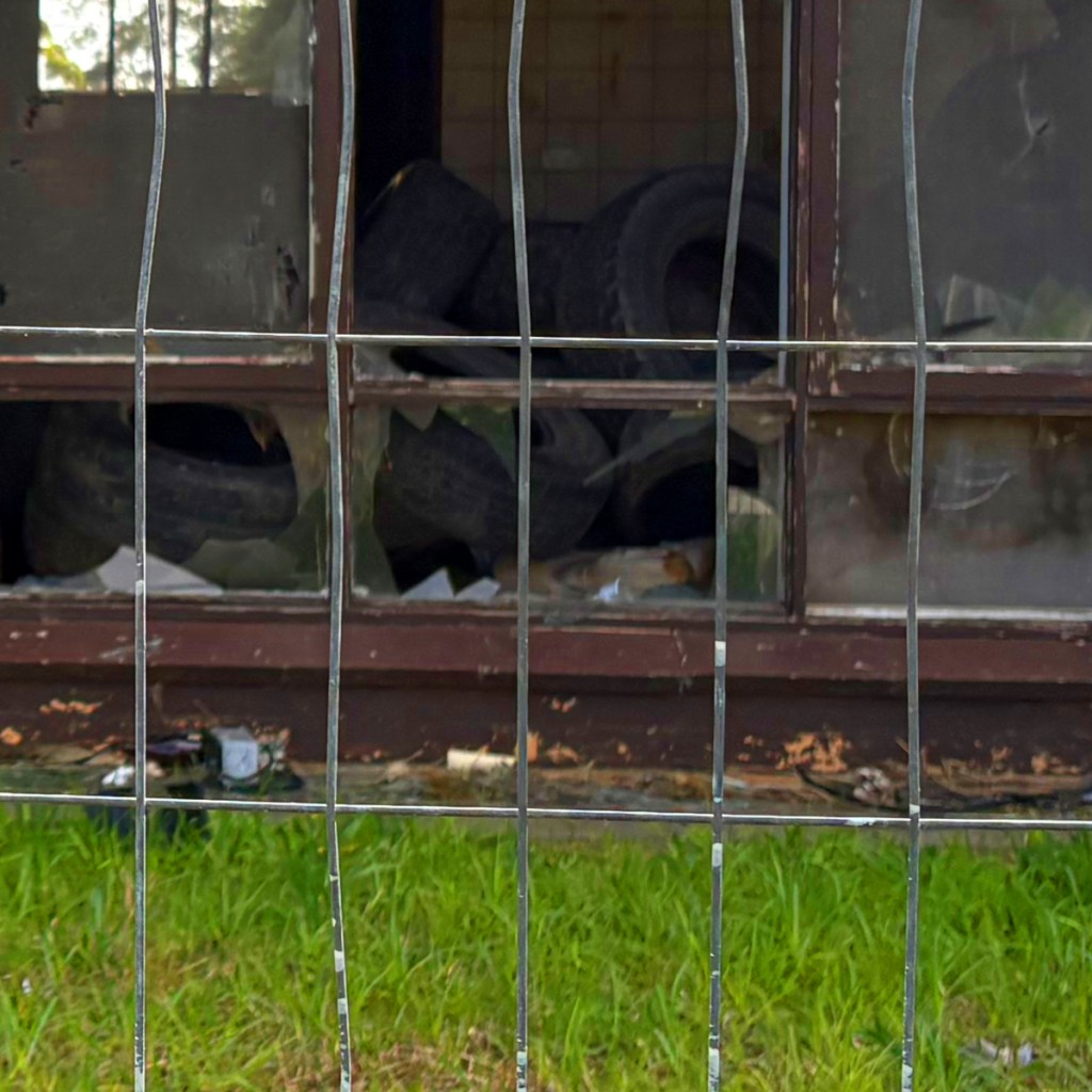 A view through a wire fence showing a broken window of an abandoned building cluttered with old tyres and debris inside.