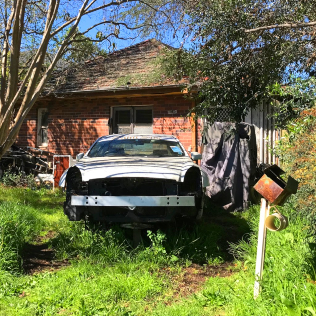 An abandoned white car without a front end, surrounded by overgrown grass in front of a red brick house. In the foreground, there is a rusty metal bird feeder.