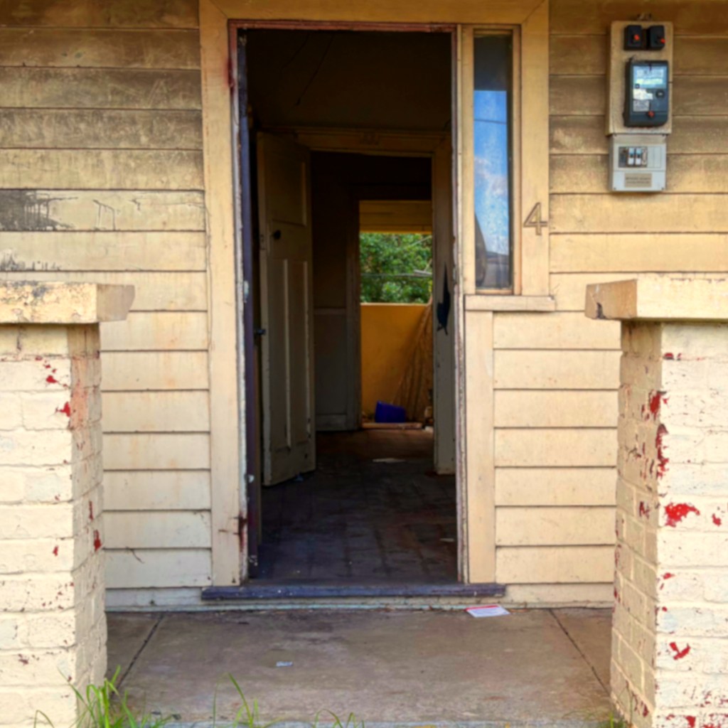 An entrance to an old house featuring a worn wooden door and faded walls, with a view of a dimly lit interior.