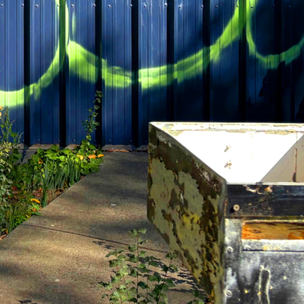 A view of a pathway lined with greenery, leading to a rusty open container against a blue backdrop with green graffiti.