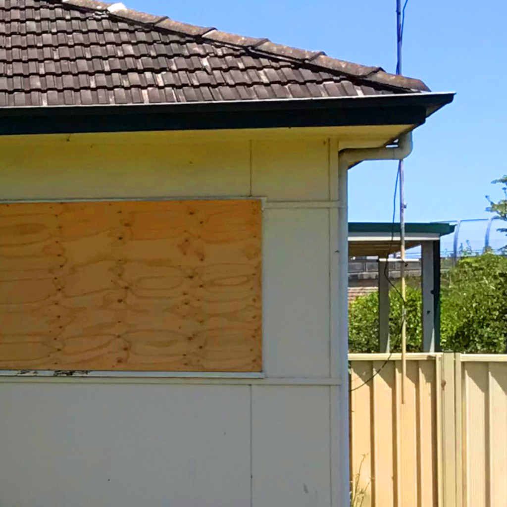 Close-up of a house exterior featuring a plywood board covering a window, accompanied by a tiled roof and clear blue sky in the background.