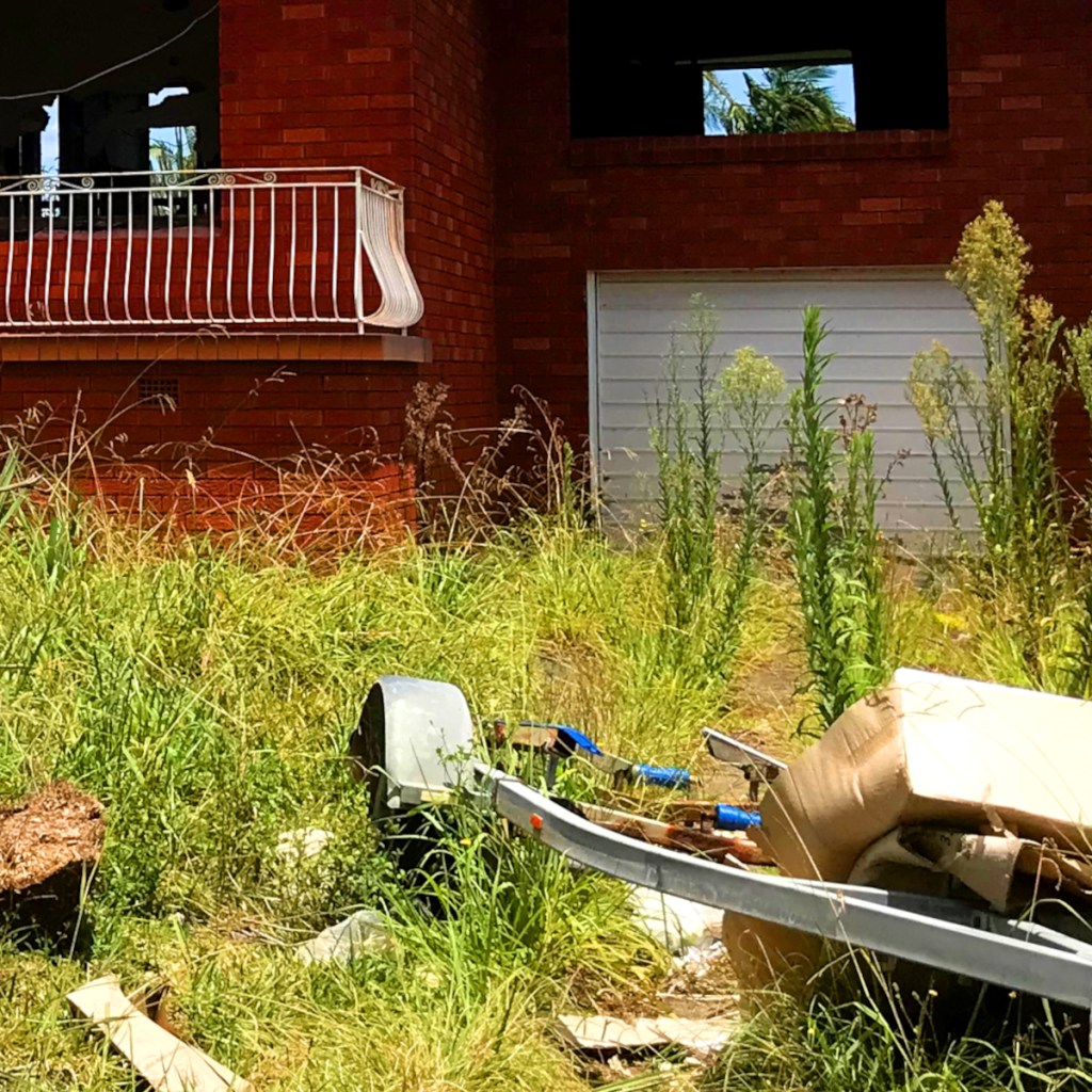 An overgrown yard in front of a brick house with a white railing balcony, featuring a discarded trailer and some rubbish among tall grass.