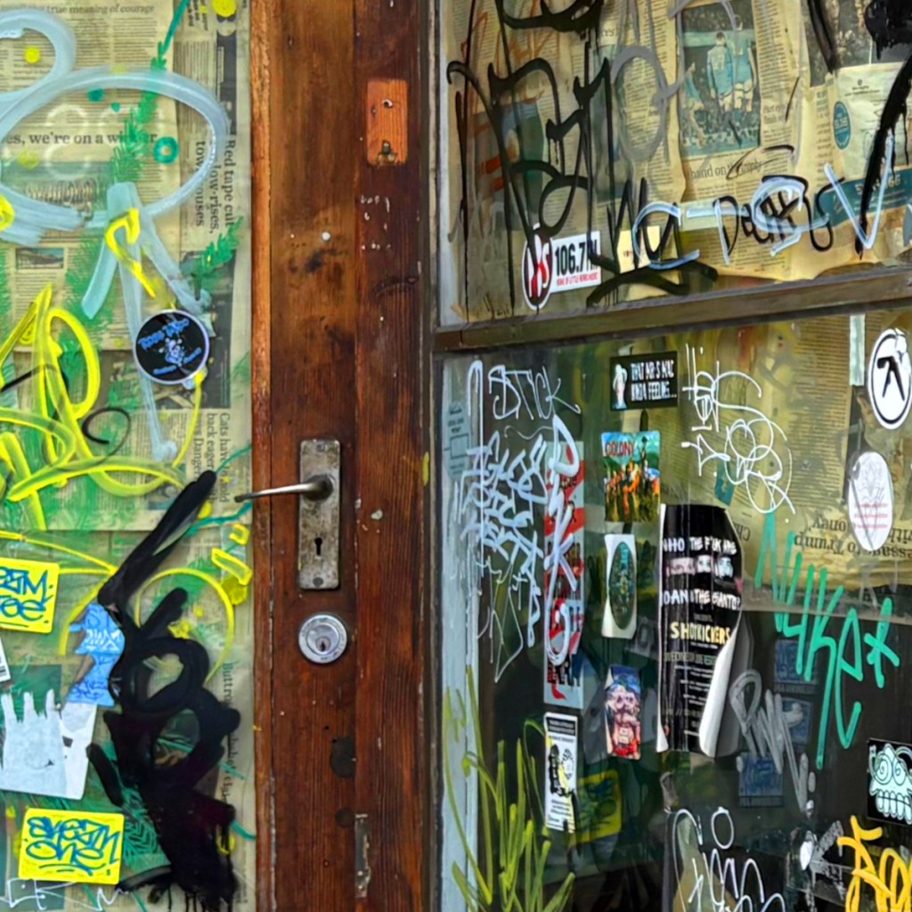 A close-up of a wooden door covered in graffiti and stickers, featuring a partly obscured glass window with newspaper clippings behind it.