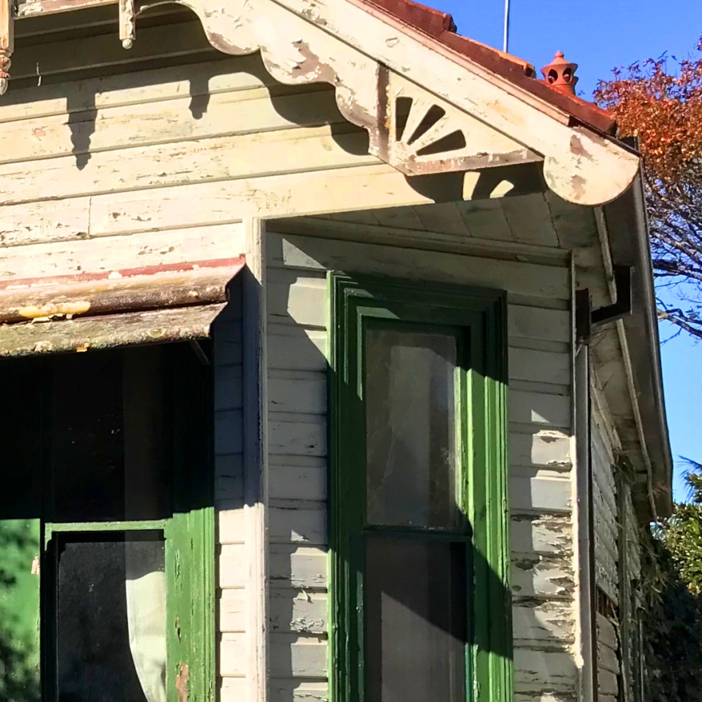 Close-up of an old wooden house corner showing weathered paint, decorative trim, and a green window frame.
